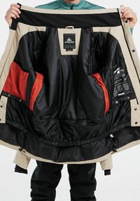 Veste beige avec une doublure noire ; comprend des poches en mesh rouge et une étiquette à l'intérieur. Tissu texturé avec des fermetures à pression et à glissière.