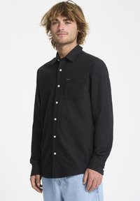 Chemise noire à manches longues, dotée d'une poche poitrine et de boutons en argent, confectionnée dans un tissu lisse et texturé.