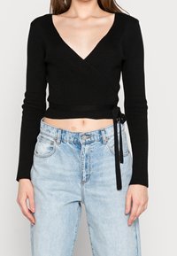 Schwarzes geripptes Wickeltop mit tiefem V-Ausschnitt, langen Ärmeln und einer Schleife in der Taille, kombiniert mit hellblauen hochtaillierten Jeans.