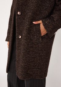 Cappotto brown texturizzato realizzato in tessuto intrecciato, caratterizzato da una vestibilità ampia, grandi bottoni e tasche laterali. Accenti di particelle dorate qua e là.