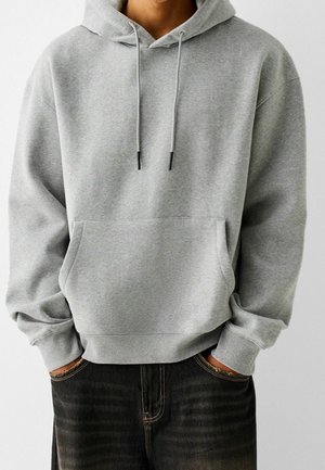 Person trägt einen hellgrauen Kapuzenpullover mit Fronttasche und Kordelzug, Hände teilweise in den Taschen von ausgewaschenen schwarzen Jeans.