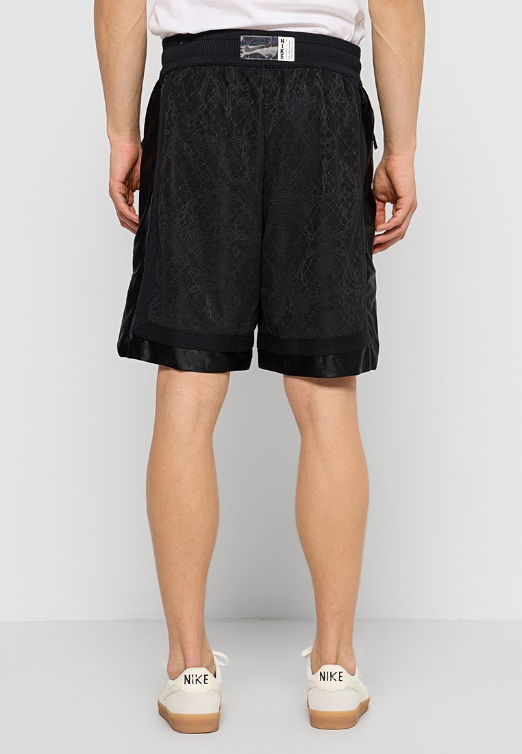 Shorts de sport noirs avec un motif texturé, taille élastique et finition en satin au bas, associés à des baskets beiges et blanches.