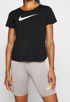 Kvinde iført sort Nike t-shirt og lysegrå Nike shorts med gult logo, stående mod en ensfarvet lys baggrund.