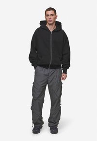 Mladý muž v čiernej mikine na zips, sivých cargo nohaviciach s veľkými vreckami a tmavých teniskách, stojí na bielom pozadí.