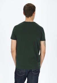 Mørkegrøn kortærmet t-shirt med rund hals. Blødt bomuldsstof. En glat tekstur, ingen synlige mønstre, båret med mørkeblå jeans.