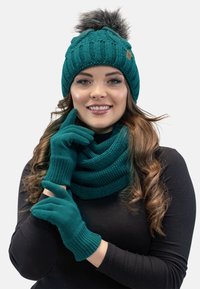 Berretto lavorato a maglia in teal con pompon in pelliccia sintetica, guanti coordinati e una sciarpa avvolta, tutti con un motivo a maglia testurizzata, indossati sopra una maglietta nera.
