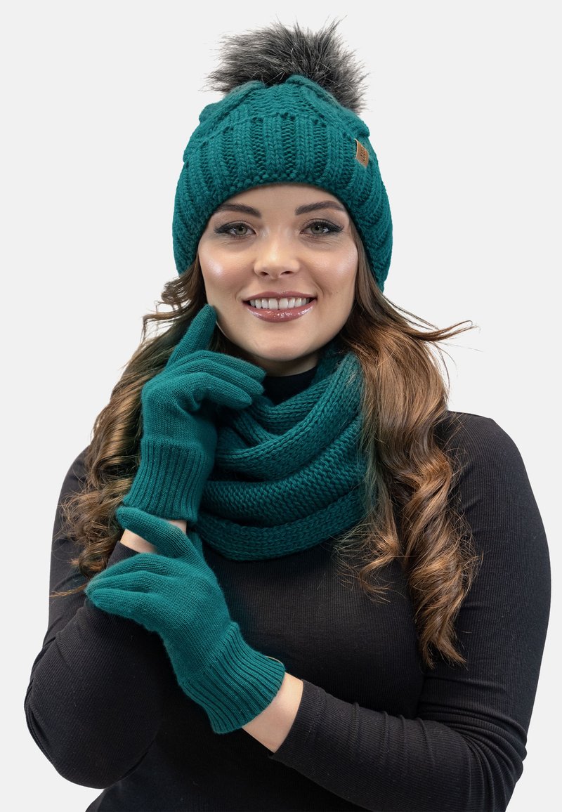 Berretto lavorato a maglia in teal con pompon in pelliccia sintetica, guanti coordinati e una sciarpa avvolta, tutti con un motivo a maglia testurizzata, indossati sopra una maglietta nera.
