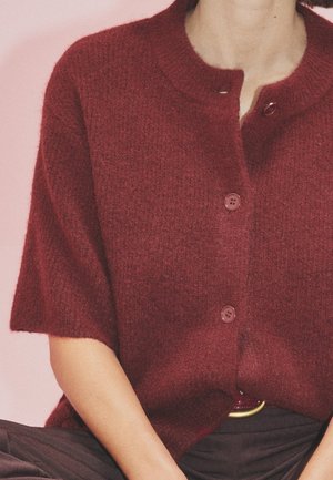 Cardigan corto bordeaux a maniche corte in tessuto morbido e testurizzato, con due bottoni e scollo rotondo. Indossato sopra pantaloni scuri con una cintura dorata.