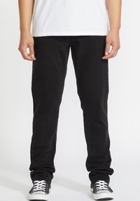 Jeans negri din denim cu un croi slim, având cinci buzunare, fermoar și o textură netedă, asortați cu pantofi din pânză negri și albi.