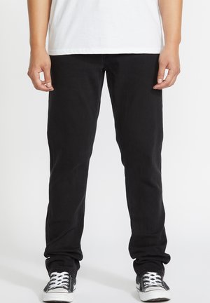 Black denim jeans with a slim fit, featuring five pockets, a zip fly, and a smooth texture, paired with black and white canvas shoes.