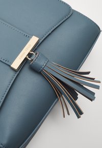 Blå handväska i konstläder med en guldfärgad platta som står "ANNA FIELD" och en läderfransdekoration. Slät textur och strukturerad design.