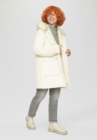 Kremowa parka z kapturem z sztucznego futra, z przednimi kieszeniami i zapięciem na zatrzaski. Noszona na zielonym golfie, zestawiona z szarymi dżinsami i beżowymi botkami.