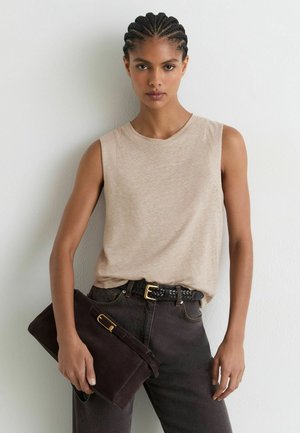 Frau mit geflochtenem Haar, die ein ärmelloses beiges Oberteil und dunkle Jeans trägt und eine große dunkelbraune Clutch vor einem einfarbigen Hintergrund hält.