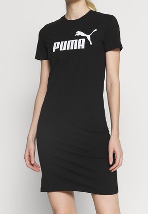 Mujer vestida con un vestido corto negro de Puma con logo blanco de Puma y texto en el pecho, de pie contra un fondo claro y liso.