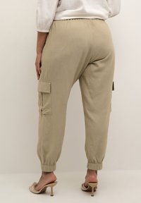 Donna che indossa pantaloni cargo beige con polsini elastici e sandali con tacco nudi, in piedi su uno sfondo chiaro e semplice.