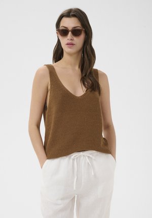 Femme portant un haut marron en maille sans manches, un pantalon blanc avec cordon et des lunettes de soleil marron, debout les mains dans les poches.