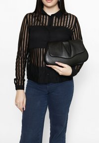 Schwarze, transparente Bluse mit langen Ärmeln, horizontalen Streifen und Kragen. Hält eine schwarze, strukturierte Clutch mit einer weichen, umschlagbaren Klappe. Denim-Jeans sichtbar.