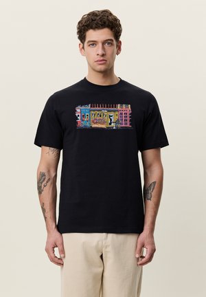 Homme aux cheveux bouclés portant un t-shirt noir avec un graphisme coloré de scène de rue et un pantalon beige clair, debout devant un fond uni.