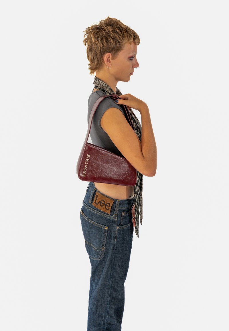 Burgundi bőr válltáska háromszög alakú, ezüst színű "EXPATRIÉ" felirattal, dombornyomva. Texturált felület és vékony pánt.