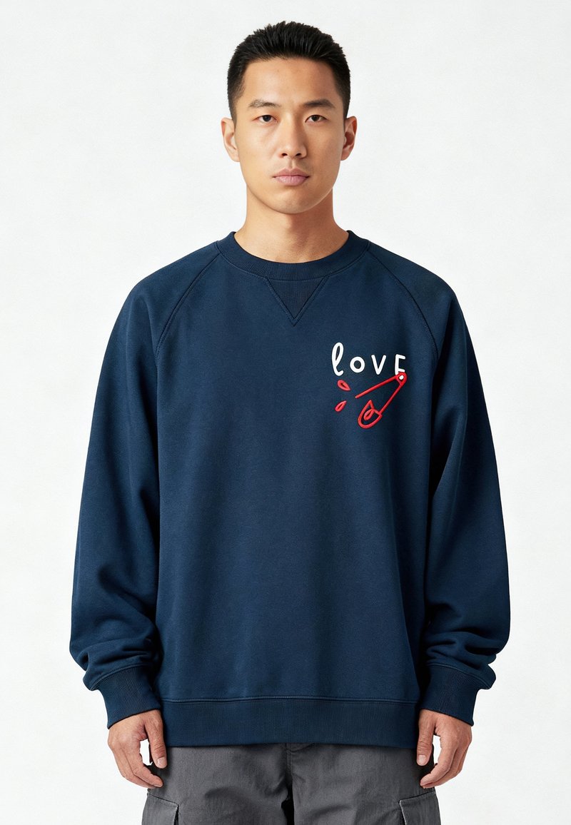 Mann trägt ein marineblaues Sweatshirt mit einer roten Sicherheitsnadel und dem Wort "Liebe" auf der Brust bestickt, steht vor einem einfarbigen Hintergrund.