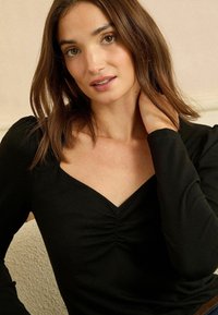 Fekete, hosszú ujjú felső négyzet alakú kivágással és középen ráncolt részlettel, sima anyagból készült. A modell az egyik kezét a nyakára helyezi.