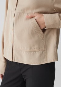 Beige jasje met een zak, gemaakt van gladde stof. Heeft een knoopsluiting aan de voorkant en een gestructureerde kraag; gedragen over zwarte broek.