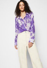 Camisa de manga longa roxa com um padrão tie-dye, com botão, e gola com corte, combinada com calças largas de cor creme com textura.