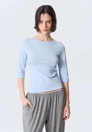 Jeune femme aux cheveux courts et foncés, portant un haut bleu clair à manches trois-quarts et un pantalon gris ample, debout avec une main dans sa poche.