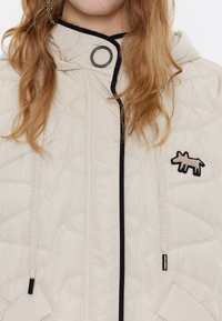 Chaqueta acolchada beige con capucha, con ribetes negros, un cierre circular y un diseño de animal bordado en el lado izquierdo.