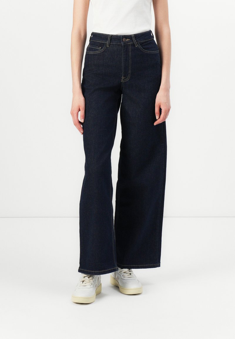 Hlače z visokim pasom in širokimi hlačnicami v temnem denimu, z petimi žepi, zlate šive in klasično zapiranje z gumbom. Oblečene z belimi športnimi čevlji.