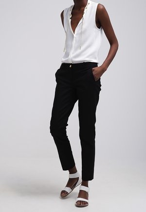 Frau trägt ärmellose weiße Bluse mit goldenen Ösen-Details, schwarze Slim-Fit-Hose und weiße Sandalen mit offenem Zehenbereich und Knöchelriemen.