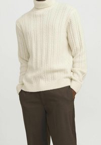 Personne portant un pull à col roulé à torsades de couleur blanc cassé, mains dans les poches d'un pantalon marron foncé, sur un fond uni.
