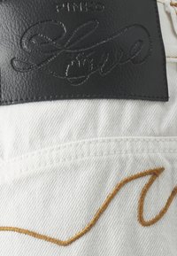 Bílý denim materiál s černou koženou štítkem s nápisem "PINCO" a "Love" ve vyraženém písmu. Obsahuje zakřivený detail žlutého stehu.