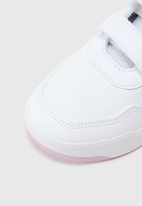 Zapatilla blanca con parte superior texturizada, punta de malla y correa ajustable. La suela tiene un sutil detalle en rosa. Forma redondeada, diseño casual.