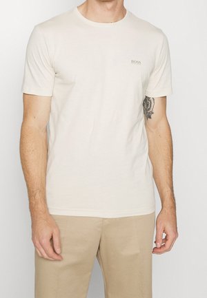 Man wearing a plain cream short-sleeve T-shirt with "BOSS" logo on left chest, beige pants, and visible floral tattoo on right arm.