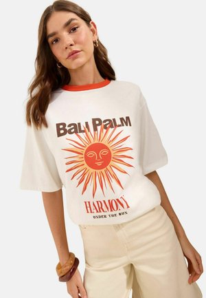 Femme portant un T-shirt blanc avec un graphique de soleil rouge et le texte "Bali Palm Harmony Under the Sun", associé à un pantalon beige clair et des bracelets.