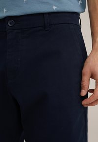 Pantalons en coton bleu marine avec une texture lisse, fermeture à bouton et poches latérales. Design ajusté montré avec une main reposant sur la taille.