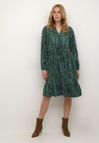 Rochie verde, cu un model, cu mâneci lungi, decolteu în V și talie cu șnur; prezintă o fustă în straturi și un material moale. Stilizată cu botine maro până la gleznă.