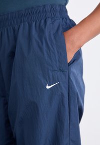 Detailní záběr osoby v námořnickomodrých šortkách Nike s elastickým pasem, s rukou zasunutou v boční kapse.