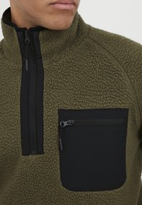 Olive green fleece pullover with a high collar, featuring a black zippered chest pocket and textured fabric for warmth and comfort.