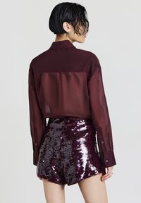 Chemise bordeaux transparente boutonnée, associée à un short taille haute en sequins violets, mettant en valeur une texture brillante et un scintillement à motifs.