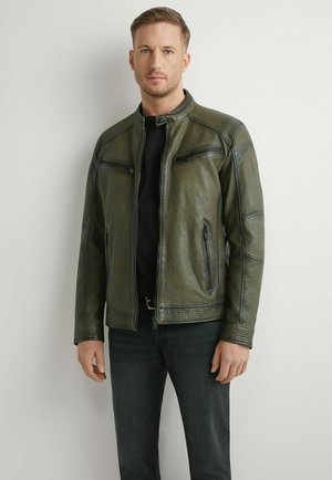 Mężczyzna w ciemnozielonej skórzanej kurtce na czarnej koszulce i ciemnych dżinsach, stojący na jasnoszarym tle.