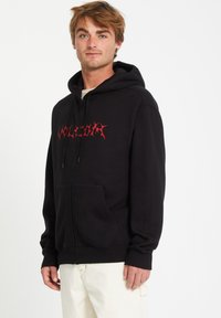 Zwarte rits-hoodie van katoen. Voorzien van een buidelzak en een rood grafisch tekstlogo op de borst. Zachte textuur en een comfortabele pasvorm.