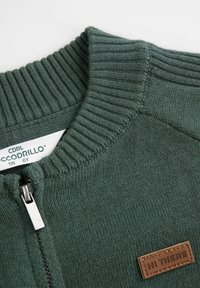 Ciemnozielony sweter w robocie z prążkowanym kołnierzem, zamkiem błyskawicznym z przodu i brązową naszywką z napisem "CZEŚĆ" blisko klatki piersiowej.