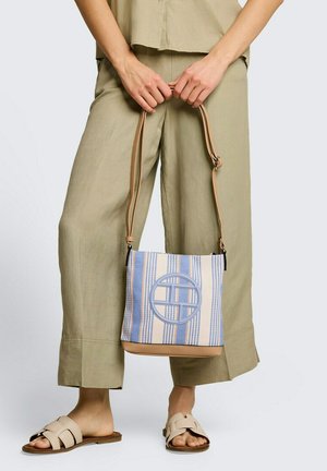 Person trägt beige Hose und Sandalen und hält eine blau-weiß gestreifte Umhängetasche mit braunem Riemen und rundem Logo.