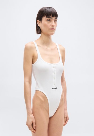 Γυναίκα με κοντά σκούρα μαλλιά που φοράει λευκό αμάνικο κορμάκι με κουμπιά και το λογότυπο "Versace" σε απλό φωτεινό φόντο.