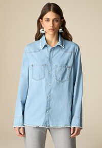 Light blue denim shirt with two front pockets, button closure, and frayed hem. Features a collar and long sleeves with a relaxed fit.