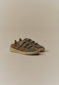 Baskets marron avec trois bandes Velcro et semelles beiges sur un fond neutre uni, légèrement orientées vers la droite.