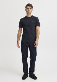 Svart bomull T-skjorte med korte ermer, med et lite rektangulært merke på brystet, kombinert med mørke denimjeans og svarte sko.