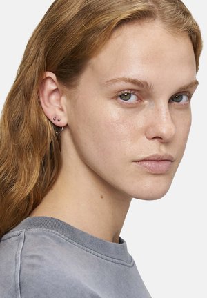 Silberne Ohrringe mit einem Ohrhänger und zwei kleinen Steckerohrringen, präsentiert an einem Modell mit welligem Haar, das einen grauen Rundhalsausschnitt trägt.